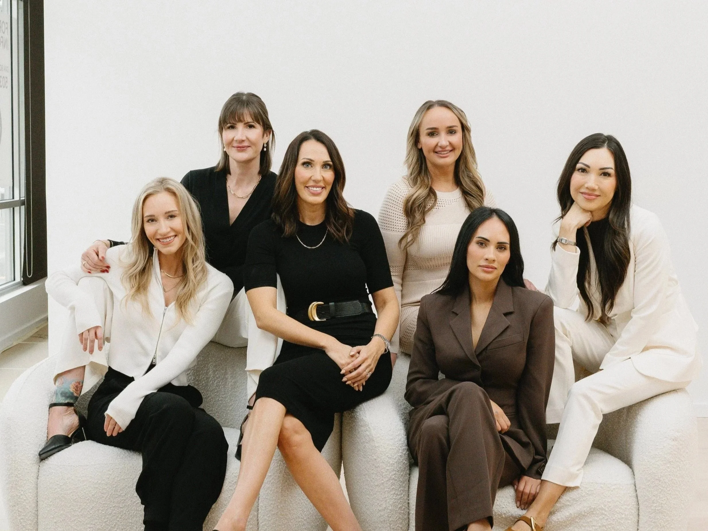 Seven women sitting and standing on a white couch in front of a plain white wall, smiling, dressed in business casual attire.
