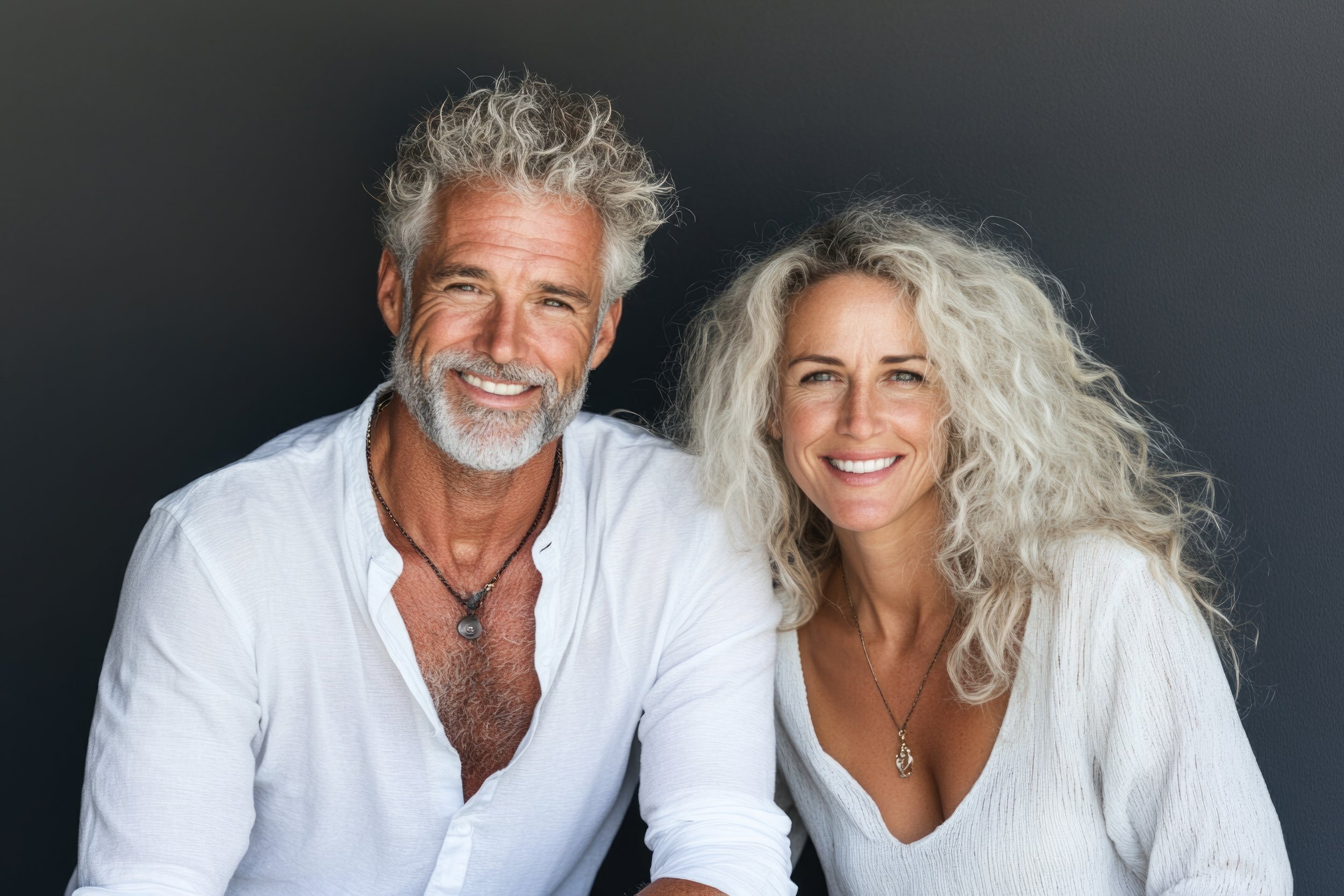 Older smiling couple with gray hair, wearing white tops, sitting close together against a dark background.