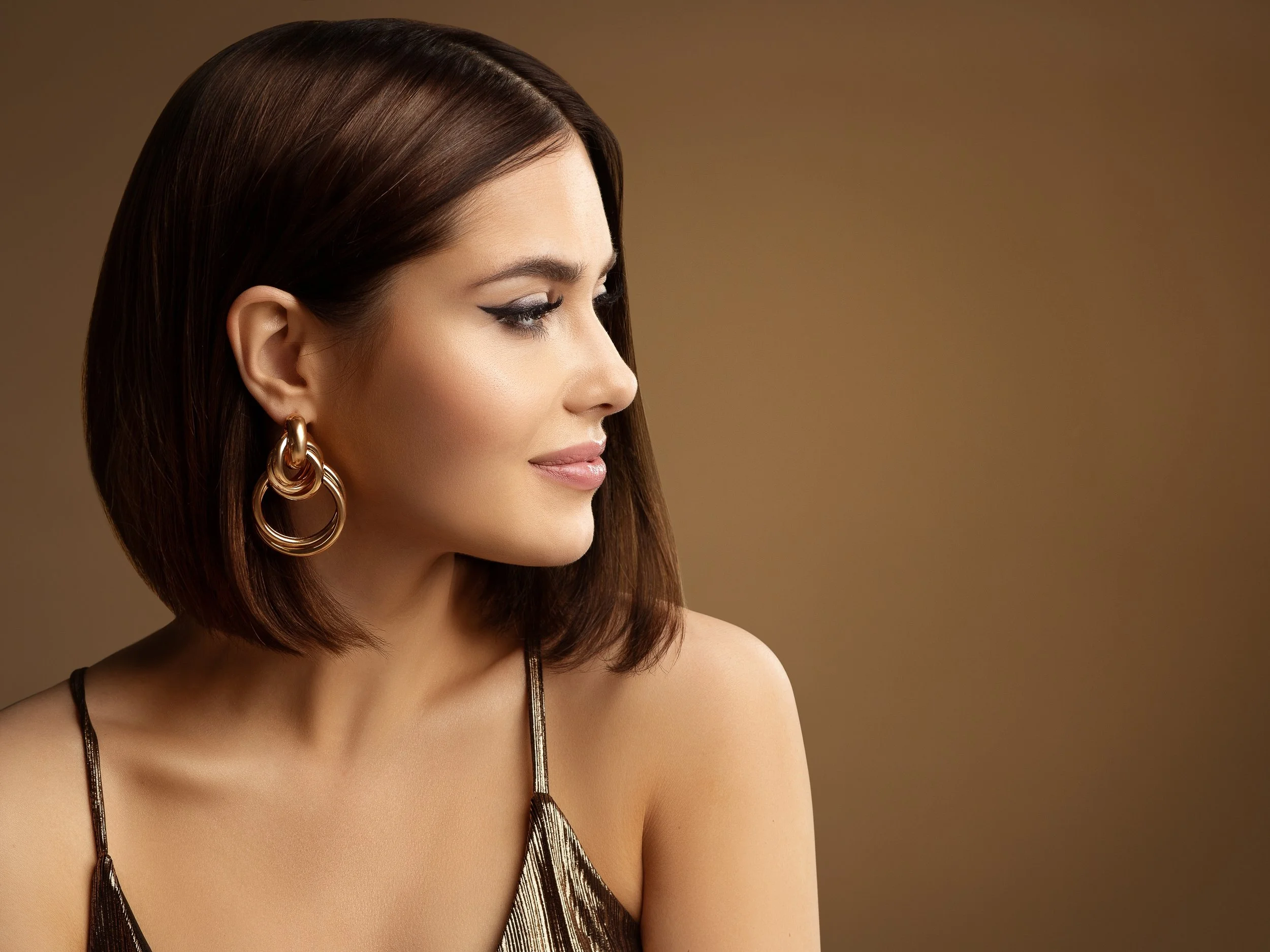 A woman with shoulder-length brown hair styled in a bob, wearing large gold hoop earrings, and a metallic spaghetti strap top, looking to the side against a brown gradient background.