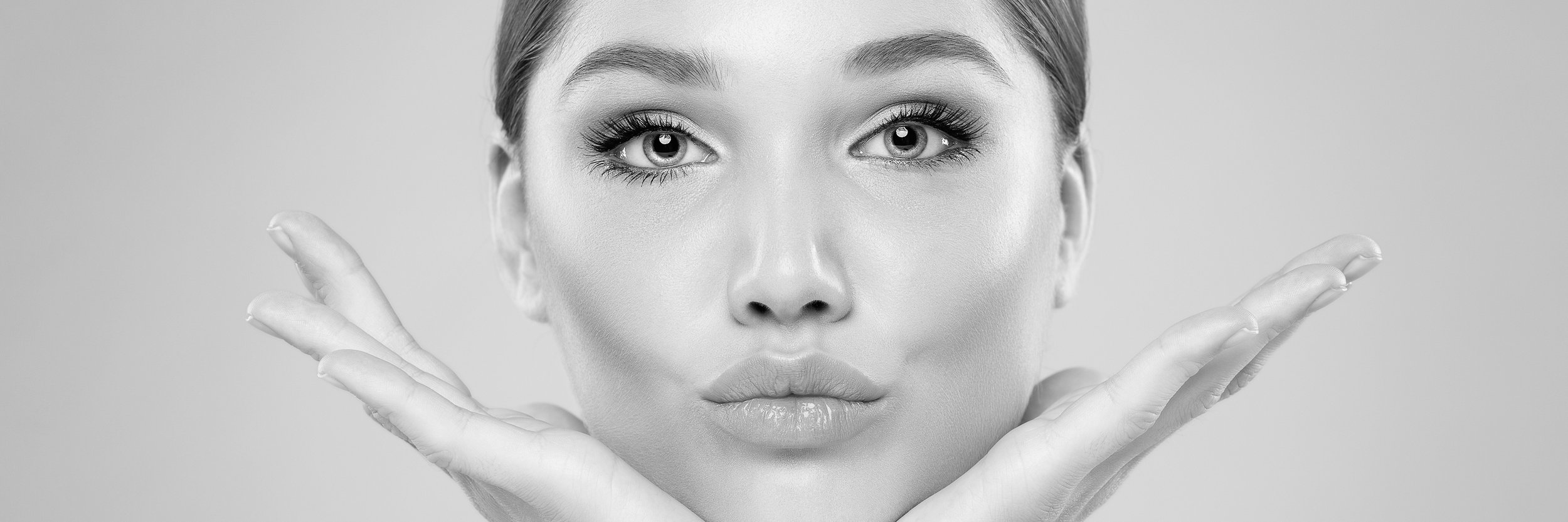 Close-up of a woman's face with makeup, touching her cheeks with her hands, in black and white.