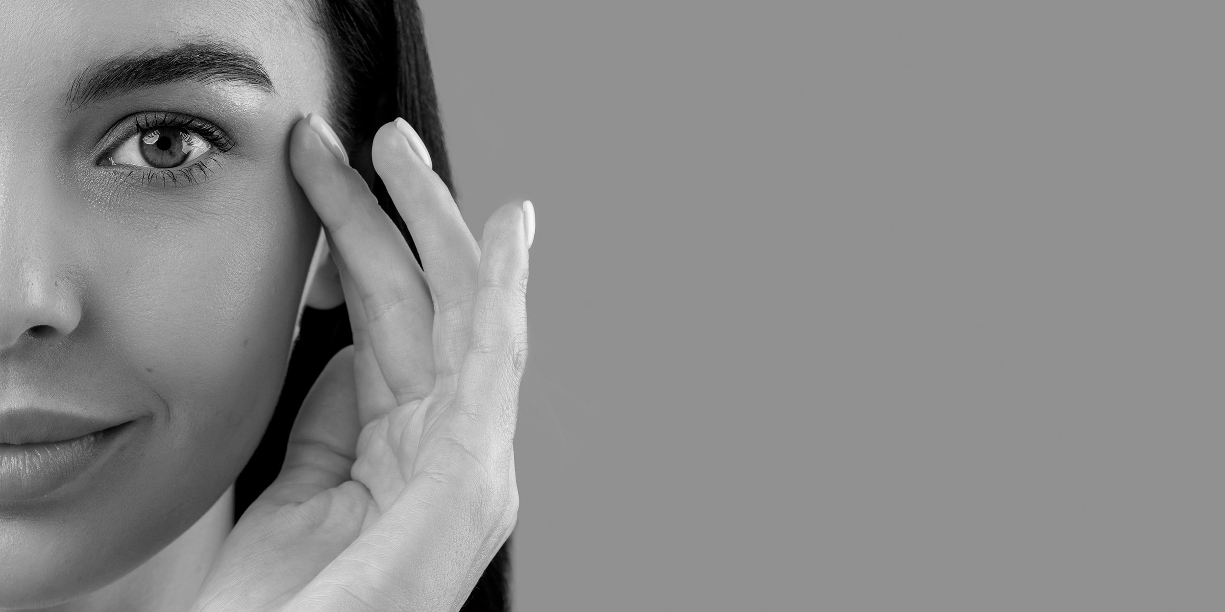 Close-up of a woman with one eye visible, touching her face near her eye with her fingers, against a plain background