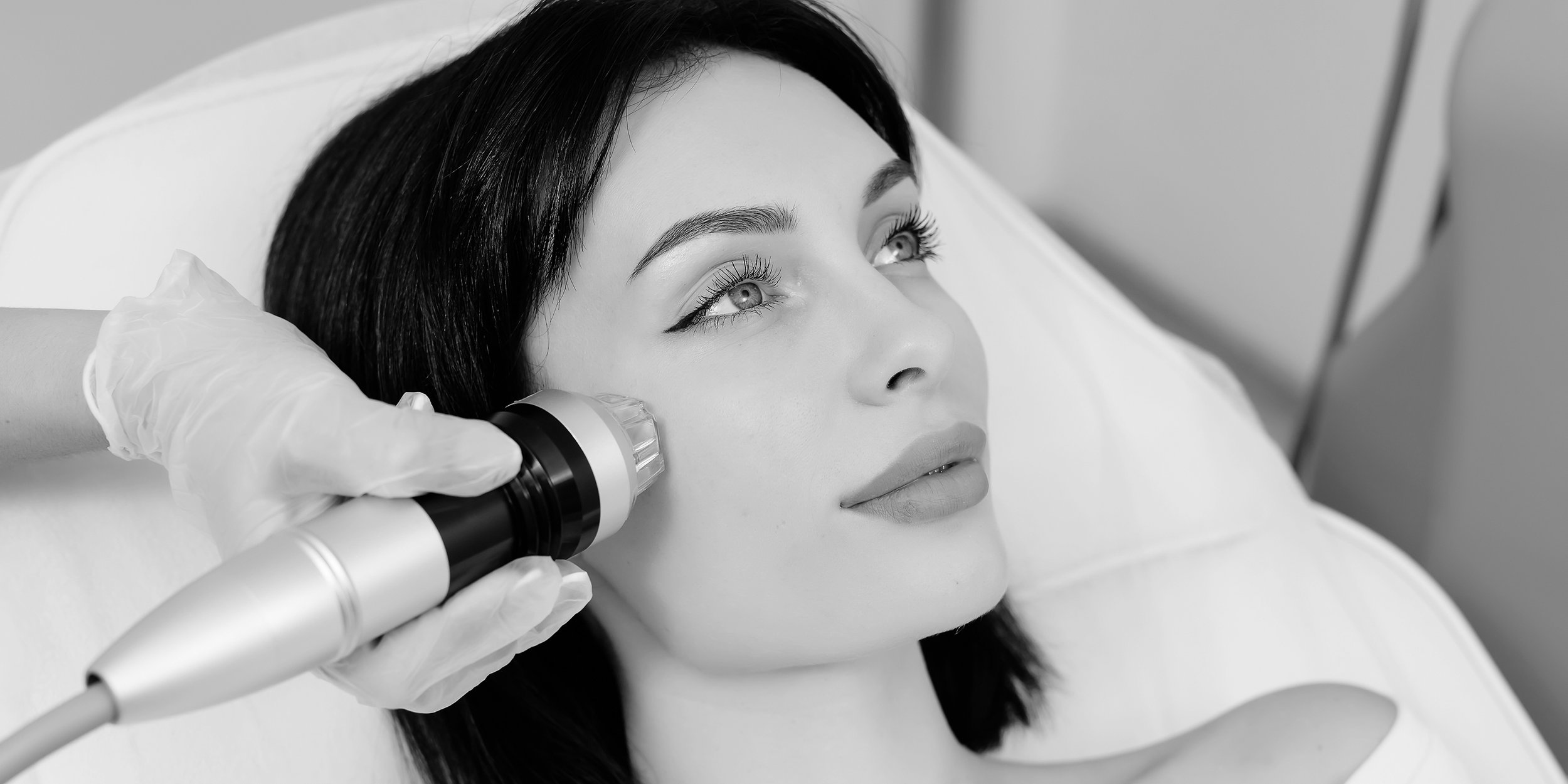 A woman lying on a medical bed receives a facial treatment with a handheld device, in a clinical setting.