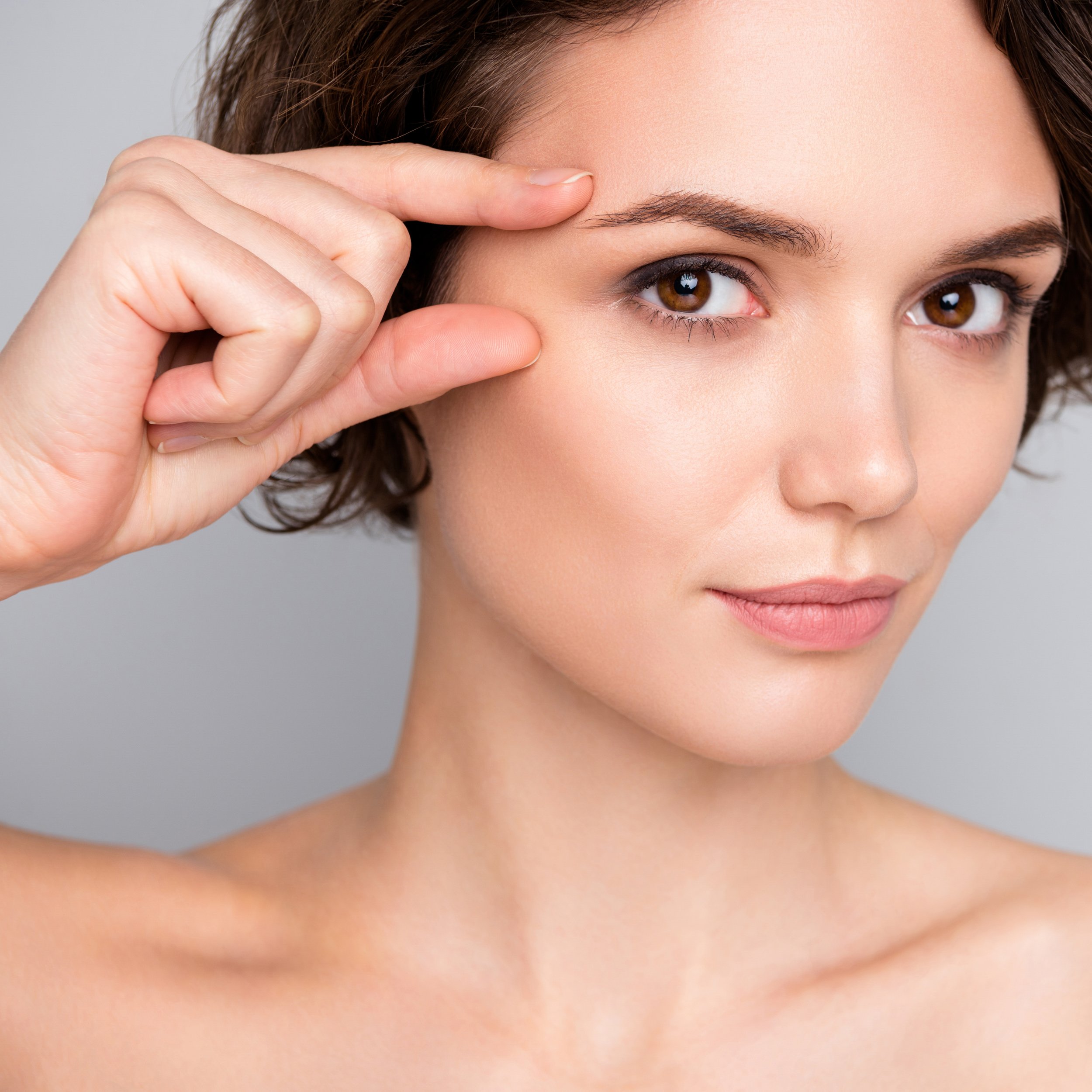 Close-up of a woman with short brown hair, touching her temple with her finger, looking directly at the camera, with smooth skin and subtle makeup.
