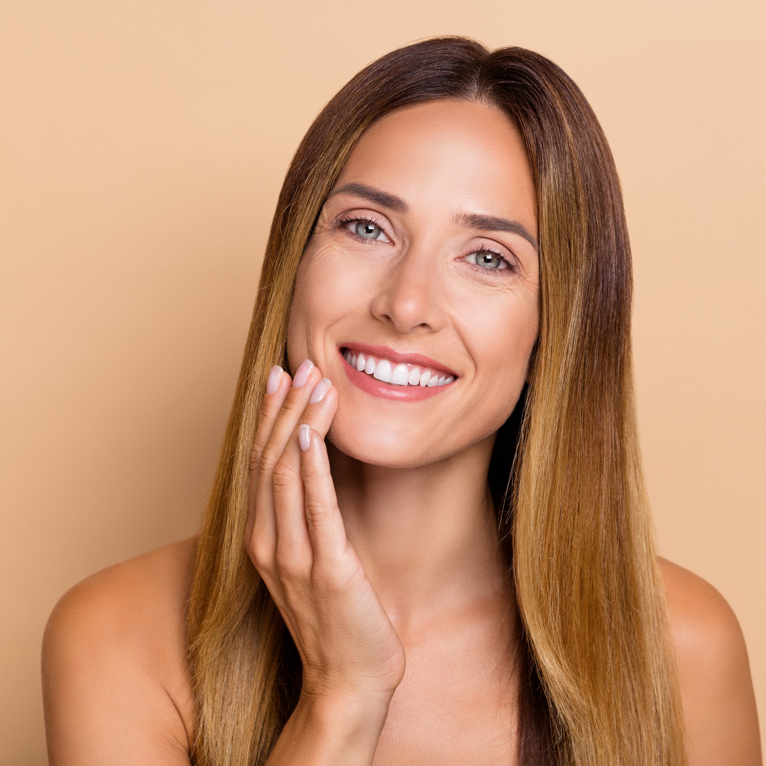 Close-up portrait of a smiling woman with long, straight brown hair, touching her face gently with her hand, against a beige background.