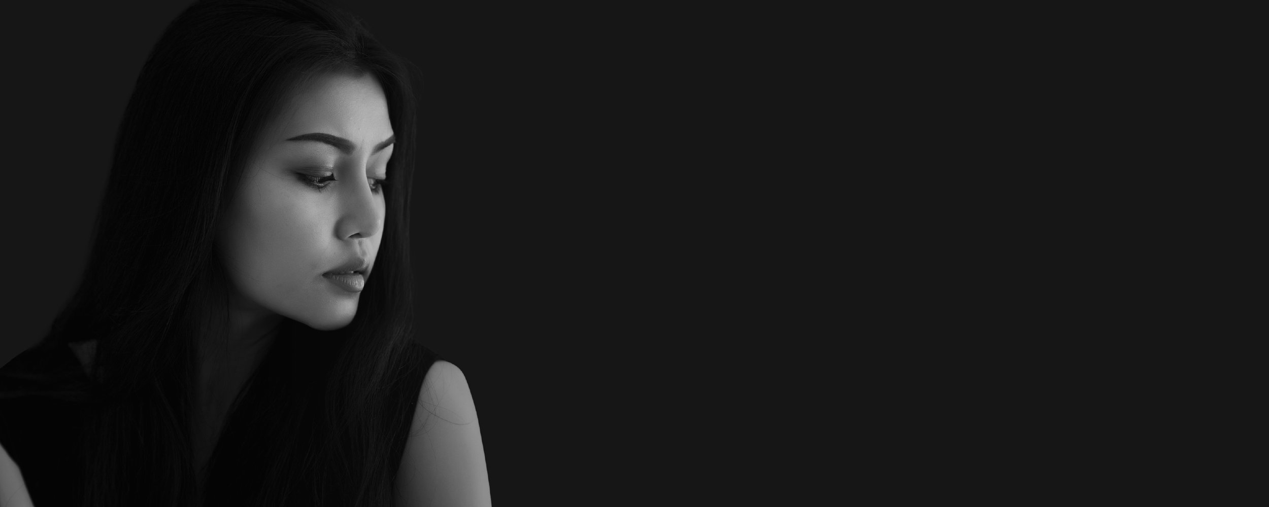 Black and white close-up portrait of a woman with long dark hair, looking down with a thoughtful expression, against a dark background.