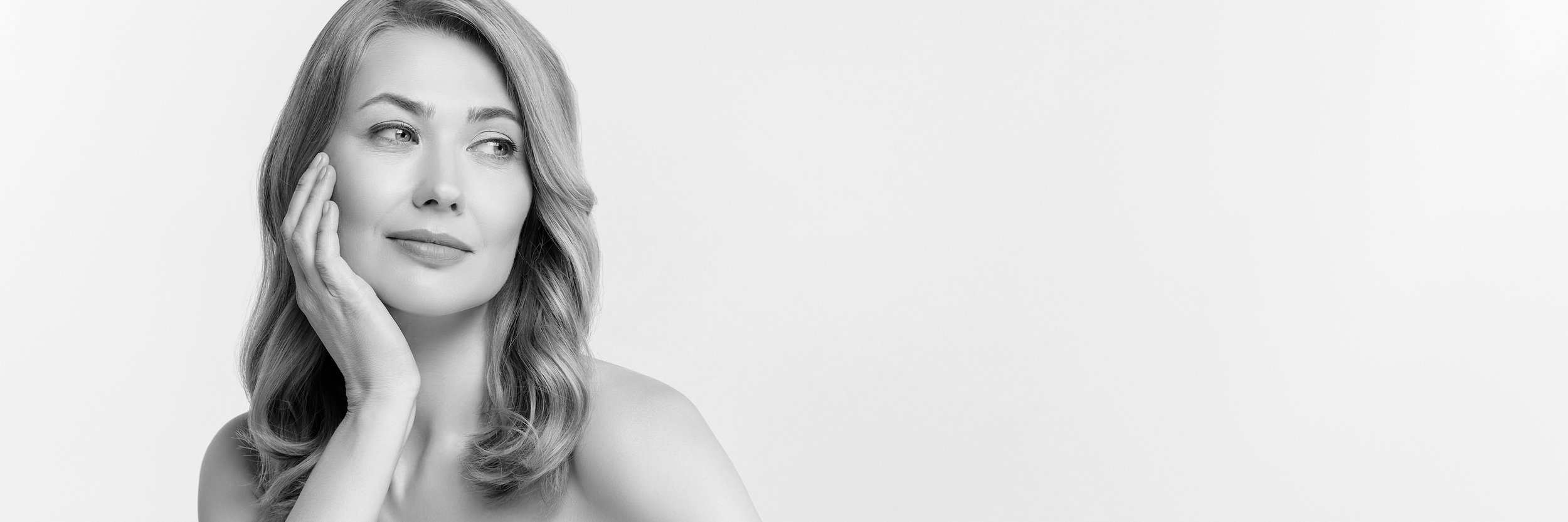 A black-and-white photo of a woman with wavy hair touching her face gently, looking slightly to the side with a calm expression, against a plain light background.