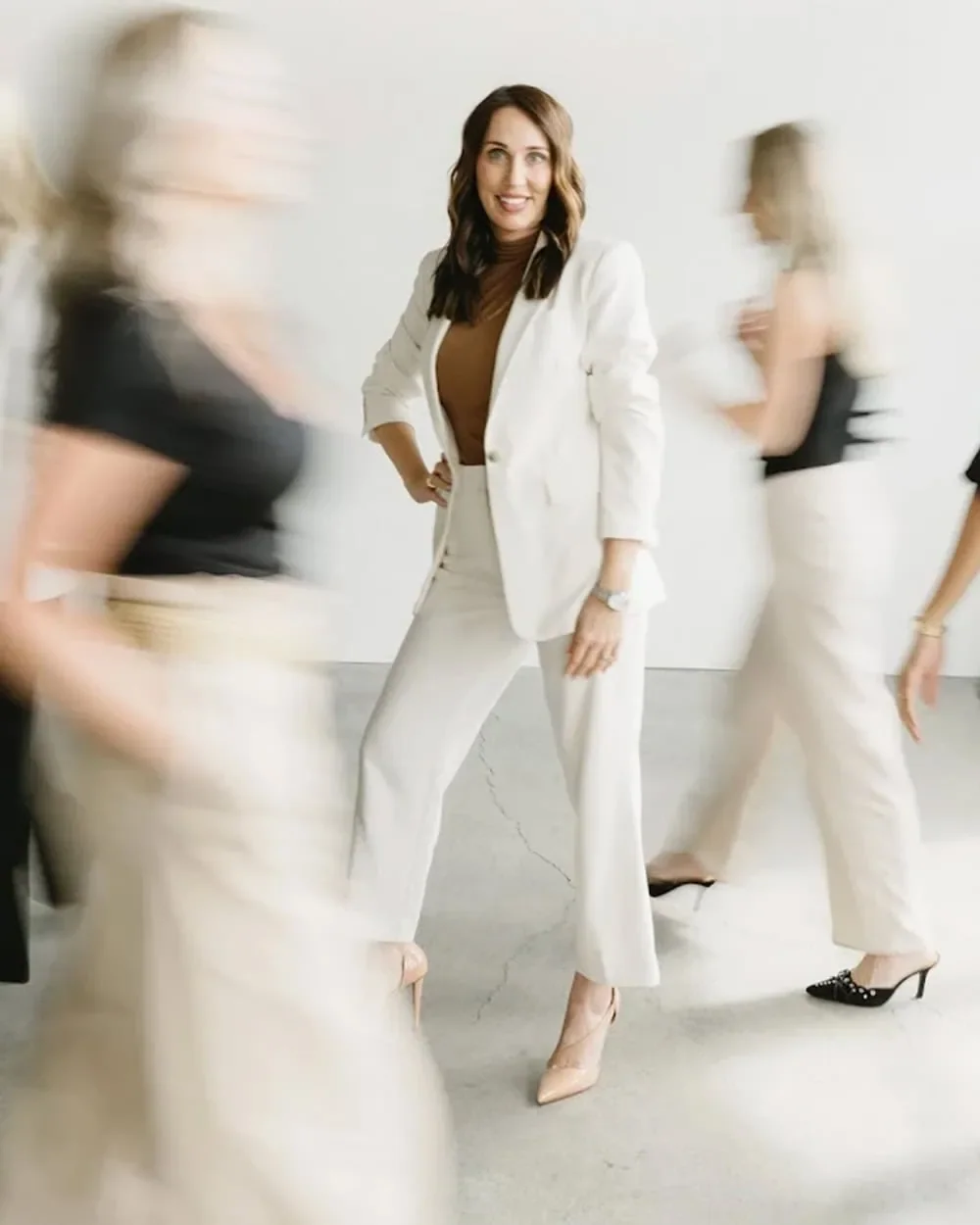Woman in white pants and blazer standing in a busy office with blurred colleagues walking past.